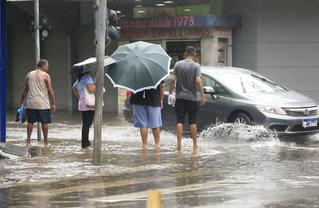Pessoas caminhando descalças em uma rua de Fortaleza alagada, algumas com guarda-chuvas, enquanto um carro passa e espirra água.