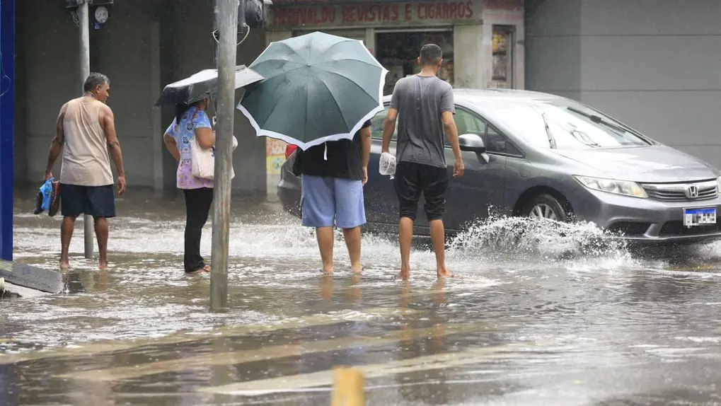 Pessoas caminhando descalças em uma rua de Fortaleza alagada, algumas com guarda-chuvas, enquanto um carro passa e espirra água.