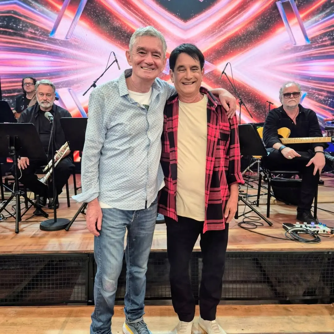 Duas pessoas estão em pé no centro de um palco iluminado, abraçadas pelos ombros. Atrás delas, há músicos sentados com instrumentos como trompete, guitarra e saxofone. O fundo exibe um painel colorido com efeitos de luz em tons de rosa, vermelho e azul, criando um visual vibrante. O piso do palco é de madeira e cabos de áudio podem ser vistos próximos aos músicos.