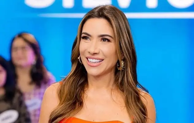 Patrícia é uma mulher branca e de cabelo castanho. Na foto, ela está no palco do programa sorrindo e usa vestido laranja.