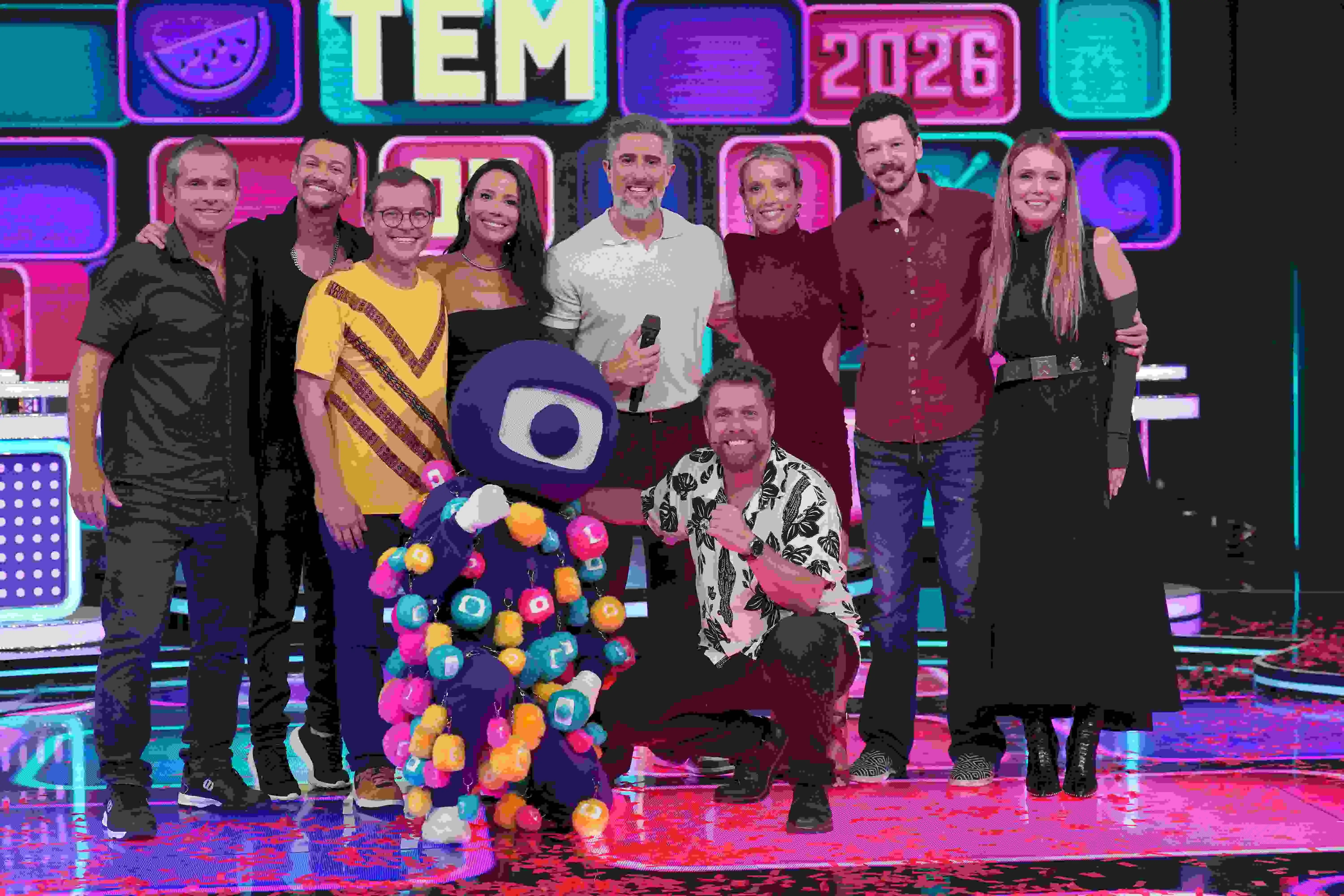 Grupo de nove pessoas, incluindo apresentadores e o mascote da Globo com ornamentos coloridos, posando em um palco de estúdio com fundo luminoso 'TEM 2026'.