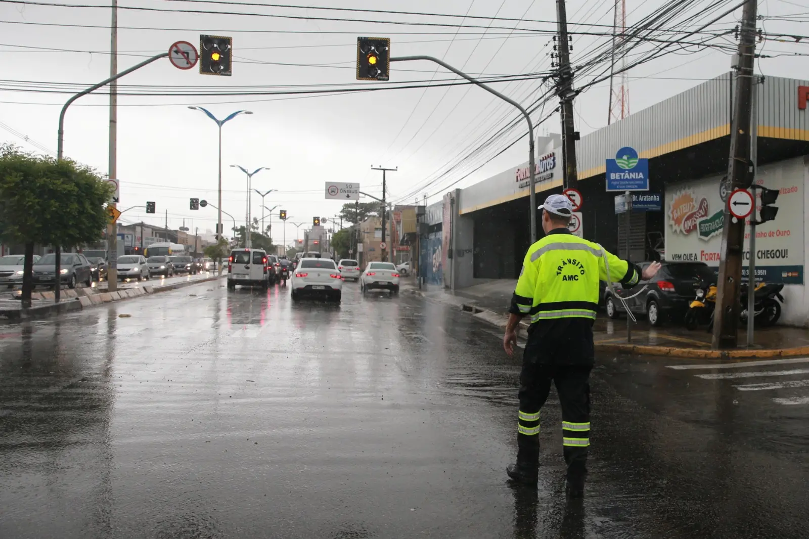 A imagem mostra um agente de trânsito da AMC (Autarquia Municipal de Trânsito e Cidadania de Fortaleza) de costas, usando um uniforme amarelo fluorescente e preto, sinalizando em um cruzamento urbano durante um dia de chuva.