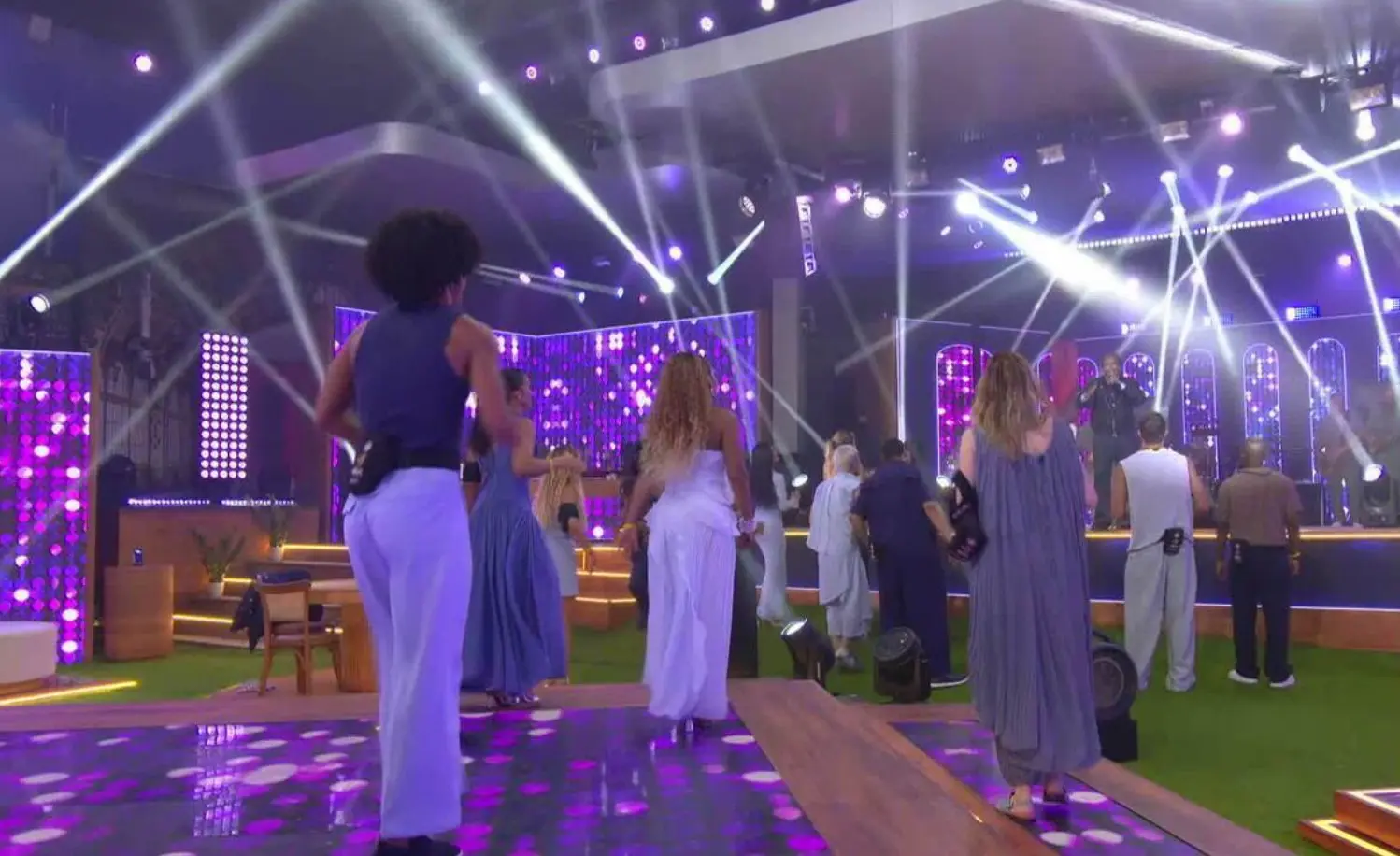 Pessoas de costas dançando em cima de um tablado luminoso e luzes iluminando o ambiente. O cantor Thiaguinho está no palco e as pessoas da plateia estão vestidas com cores azuis.