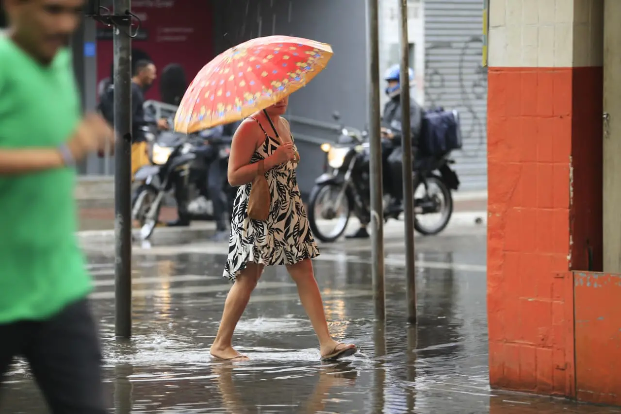Uma mulher caminha sob chuva intensa em uma rua alagada, protegendo-se com um guarda-chuva estampado em tons de vermelho e laranja. Ela veste um vestido curto de folhagens em preto e branco e chinelos, enquanto ao fundo, motociclistas aguardam no asfalto molhado que reflete a luz da cidade.