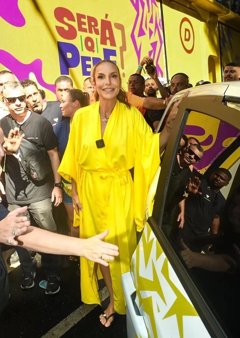 Mulher com cabelo preso em rabo de cavalo trajando um casaco amarelo e sorrindo. Ao lado, pessoas tentam abrir passagem para entrada dela em um carro preto.