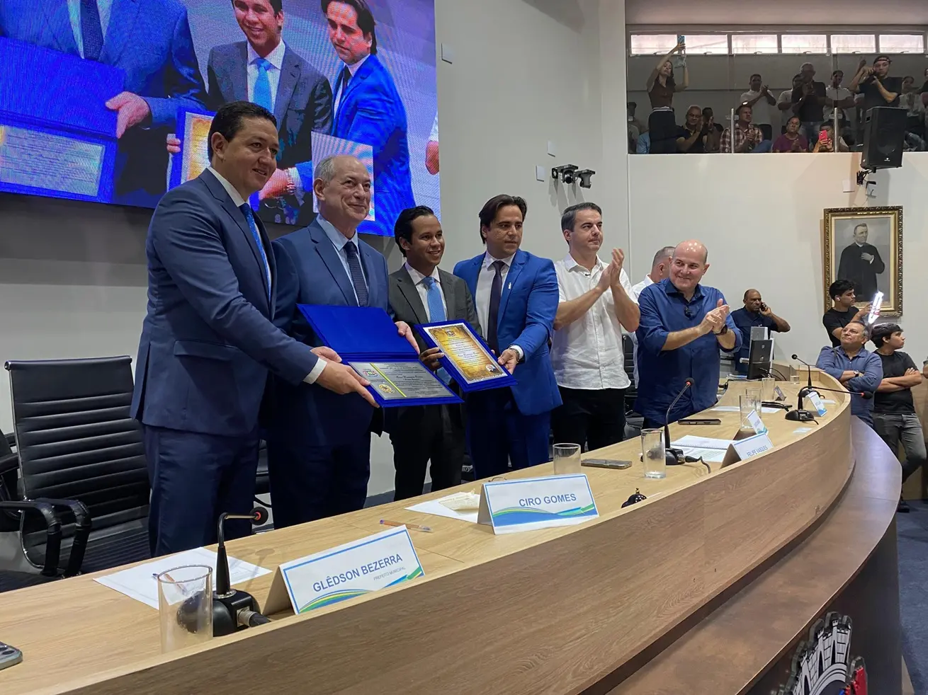Ciro Gomes e Glêdson Bezerra seguram placa de homenagem azul em evento solene, cercados por aliados e aplausos em um auditório lotado.