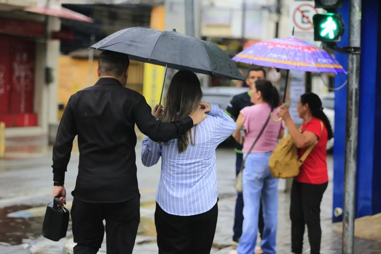 Pessoas atravessam uma rua em dia chuvoso. Em primeiro plano, um casal visto de costas caminha junto sob um guarda-chuva preto, com o braço de uma pessoa apoiado no ombro da outra. Ao fundo, outros pedestres seguram guarda-chuvas coloridos, e o semáforo de pedestres está verde.