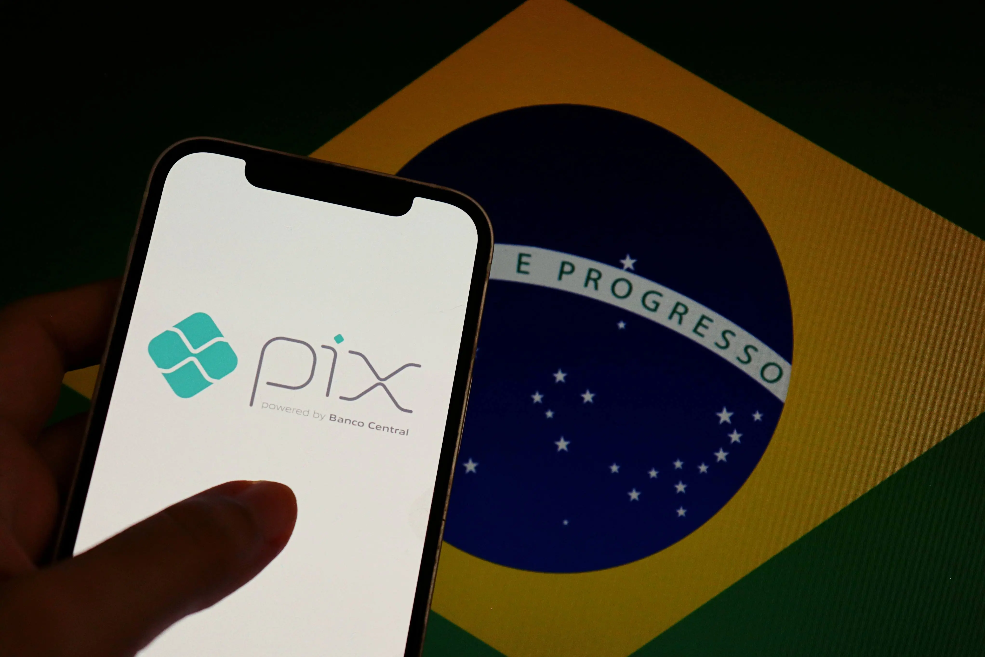 Imagem de um celular com o nome e símbolo do pix. Atrás do celular tem uma bandeira do Brasil.