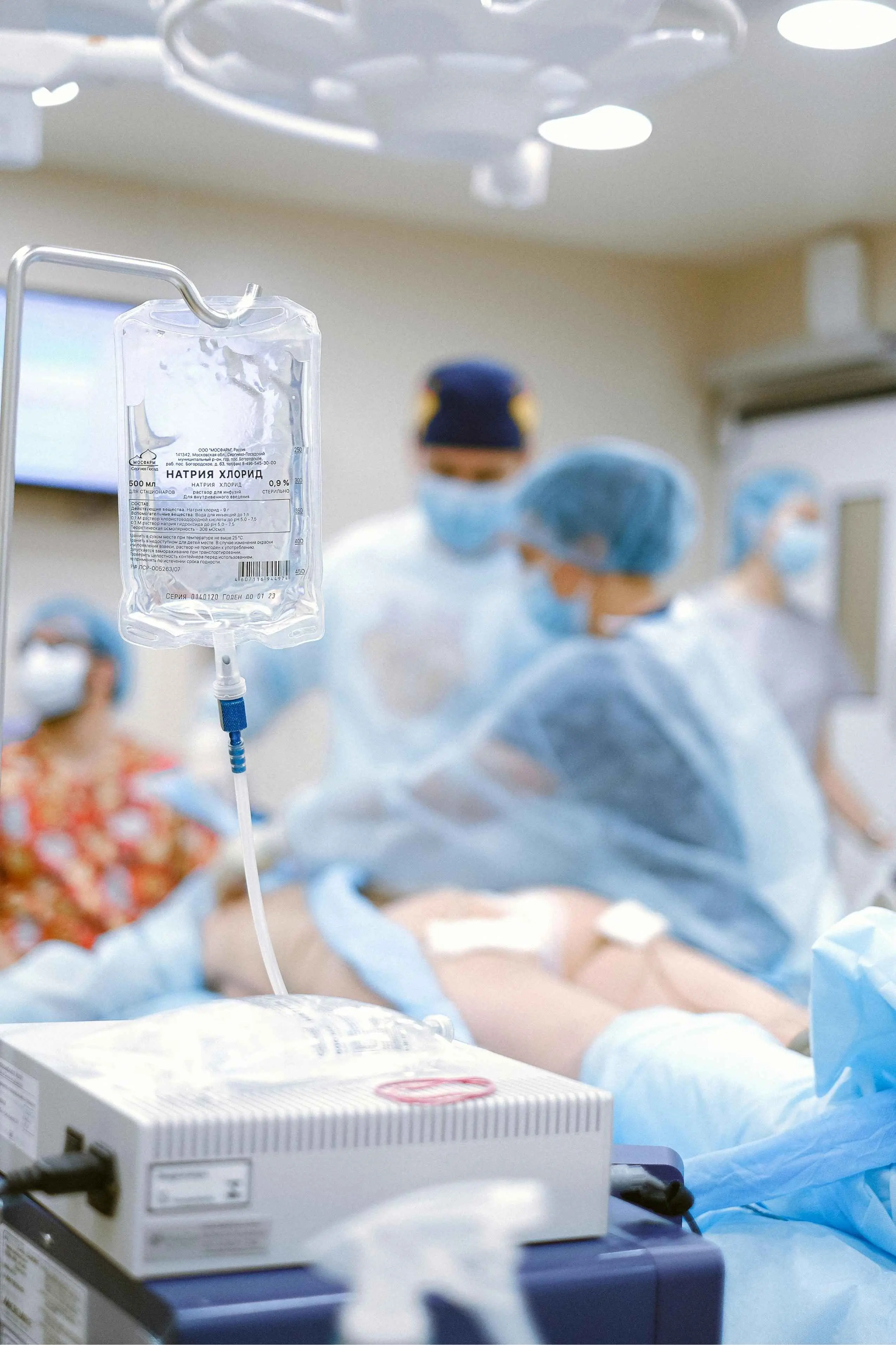Bolsa de soro pendurada em suporte em primeiro plano; ao fundo, desfocados, profissionais de saúde com máscaras e aventais realizam um procedimento cirúrgico em uma sala de cirurgia iluminada.