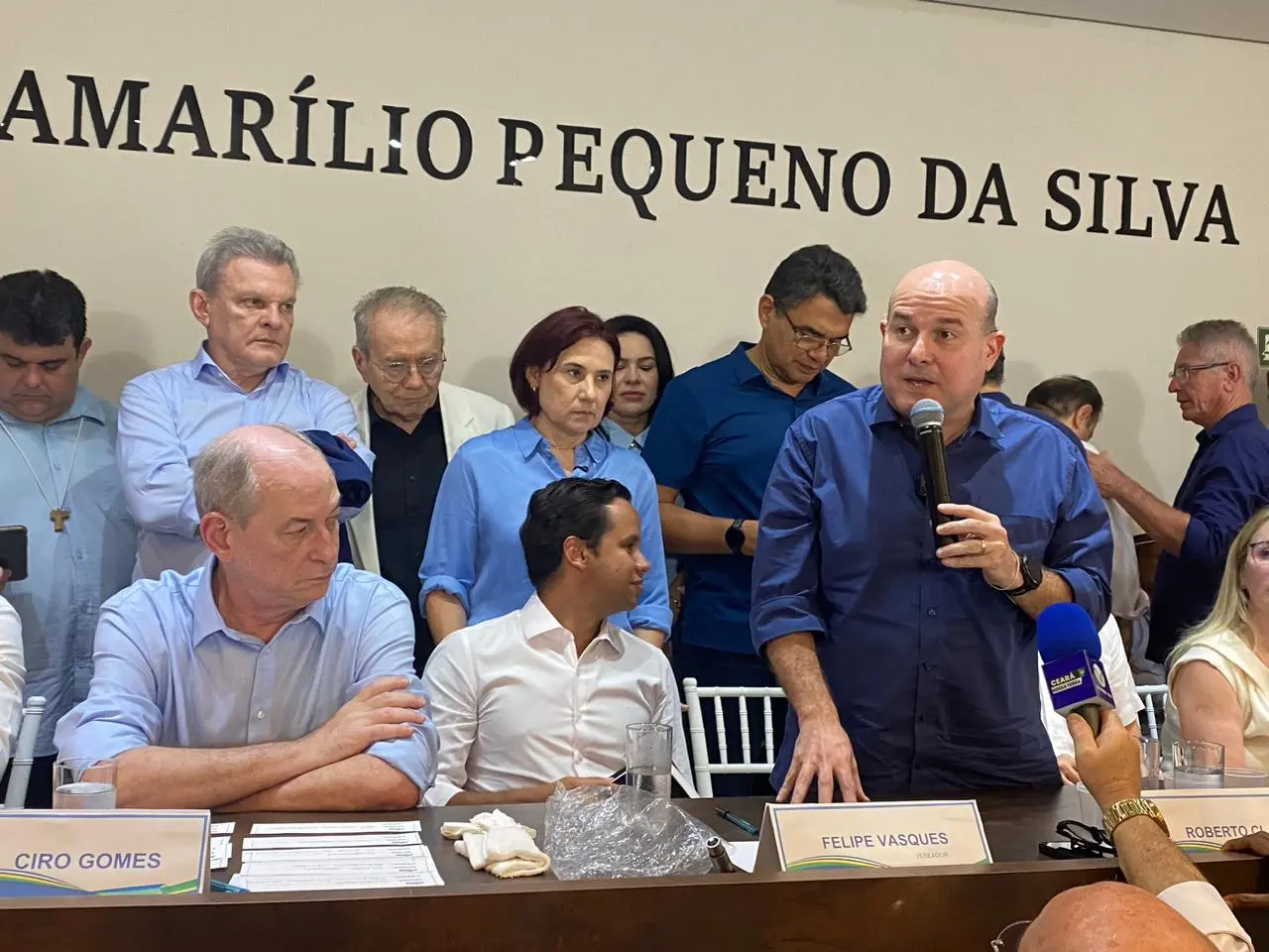 Homem de camisa azul fala ao microfone durante reunião política, em uma mesa com outros participantes sentados e em pé ao fundo. À frente, placas com nomes como “Ciro Gomes” e “Felipe Vasques”. Um repórter segura um microfone com logomarca de TV. Na parede atrás do grupo, lê-se “Amarílio Pequeno da Silva”. Ambiente interno, clima formal.