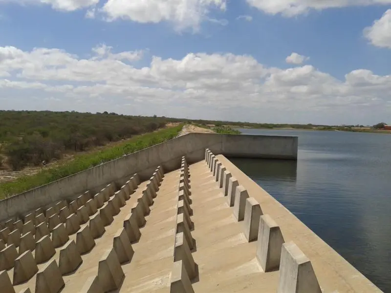Uma estrutura de concreto com blocos escalonados compõe o vertedouro do Açude Curral Velho, em Morada Nova, sob um céu azul ensolarado com nuvens esparsas. A água calma do reservatório se estende à direita, contrastando com a vegetação típica da caatinga que margeia a barragem ao fundo.