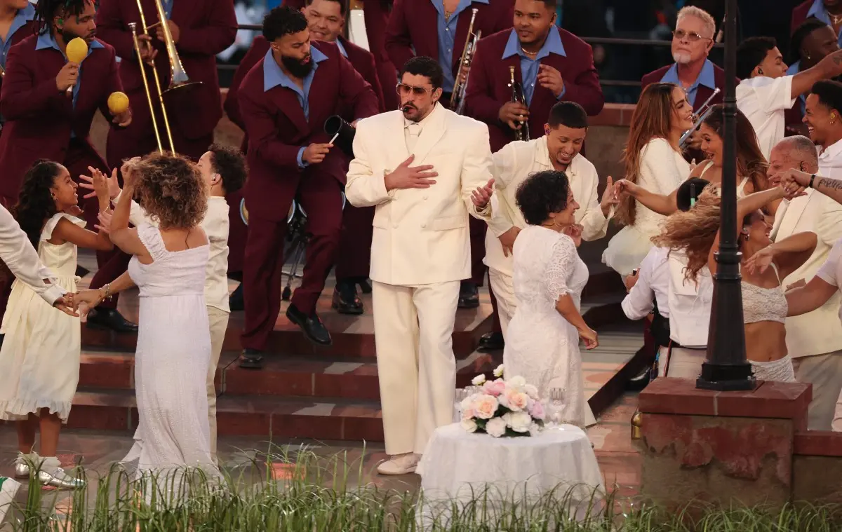 Imagem de uma celebração ao ar livre com o cantor Bad Bunny e pessoas dançando e tocando instrumentos musicais, incluindo crianças e adultos, em um ambiente festivo.