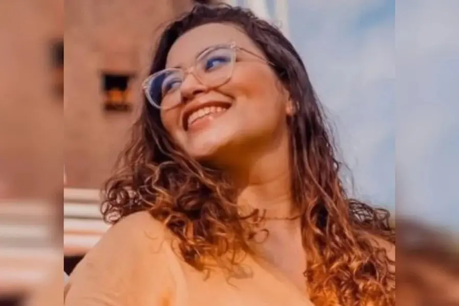 Mulher sorridente com cabelos cacheados e óculos de armação transparente em um dia ensolarado, cenário urbano ao fundo.