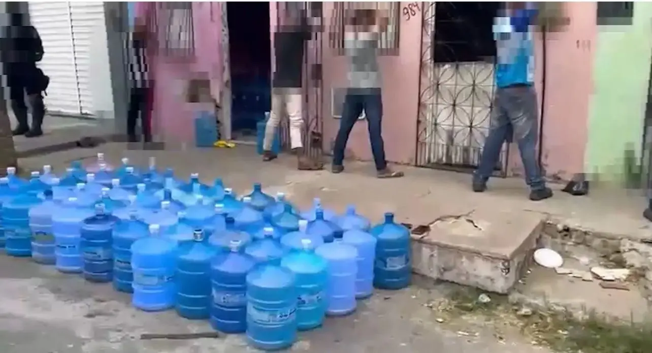 homens de costas maos para o alto na parede garrafoes de agua na calcada.