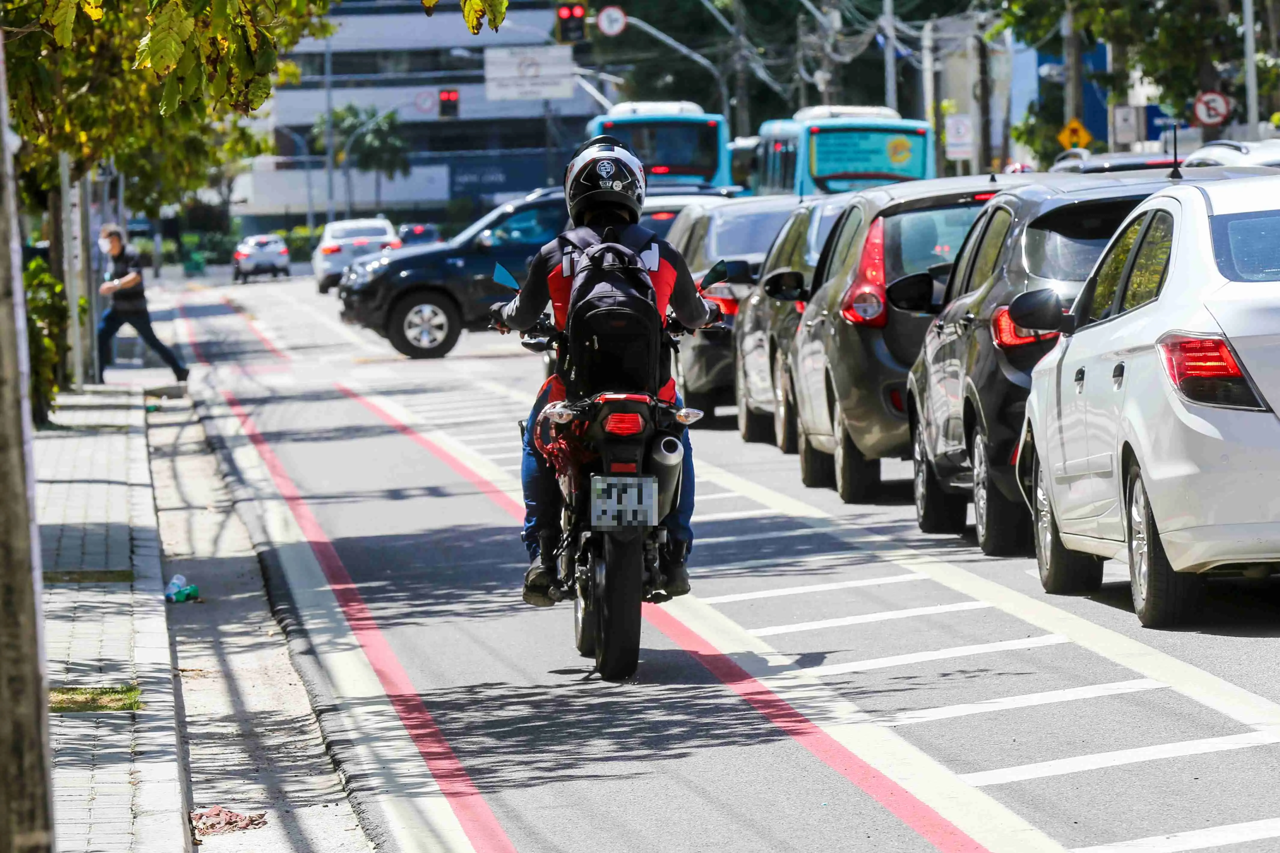 Motociclista com mochila e capacete em movimento em uma faixa exclusiva vermelha, ao lado de uma fila de carros parados em congestionamento urbano.