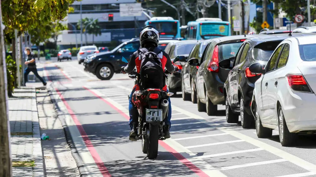 Motociclista com mochila e capacete em movimento em uma faixa exclusiva vermelha, ao lado de uma fila de carros parados em congestionamento urbano.
