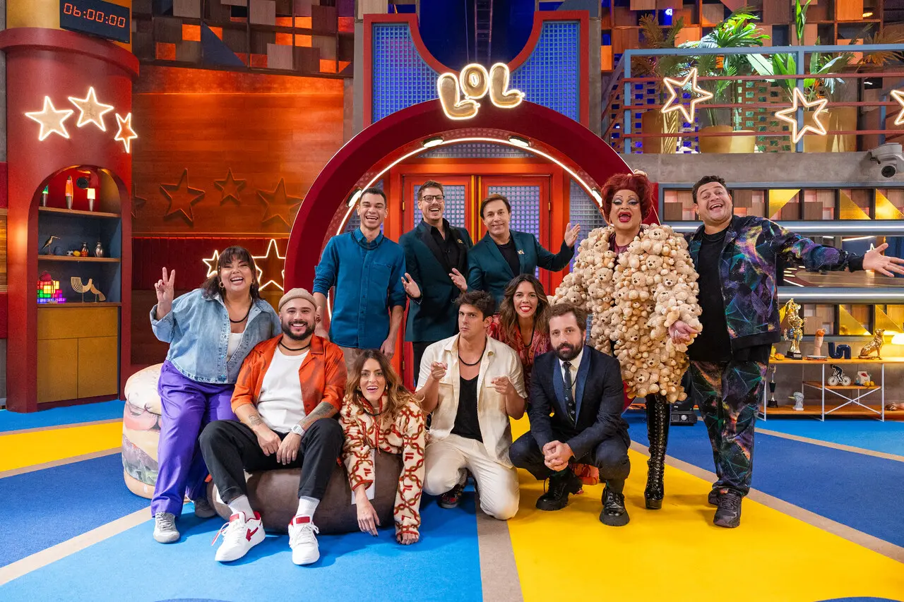 Grupo de participantes posando no cenário colorido de “LOL”, diante de um arco vermelho iluminado, com figurinos vibrantes e elementos decorativos divertidos ao redor.