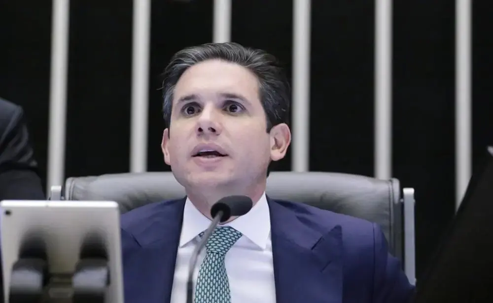 Homem de terno e gravata verde falando em microfone em um plenário.