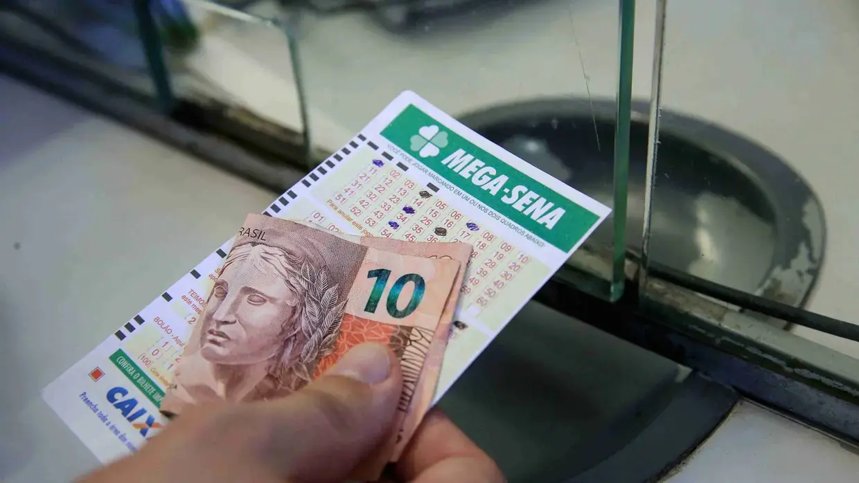 foto mostra mão segurando cartela da Mega Sena e uma nota de R$ 10 reais.
