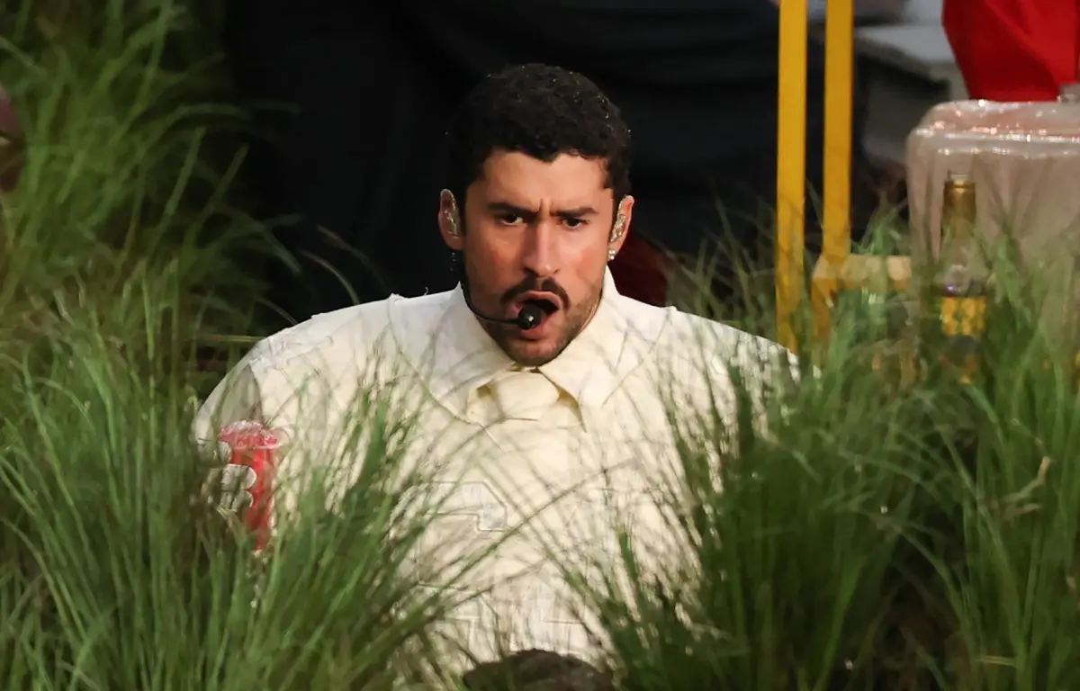 O cantor Bad Bunny, com expressão intensa e boca aberta, aparece em meio a uma densa vegetação de grama alta e verde durante sua performance no show de intervalo do Super Bowl. Ele veste uma camisa branca com gravata combinando e utiliza um microfone headset, enquanto segura um objeto vermelho na mão direita.