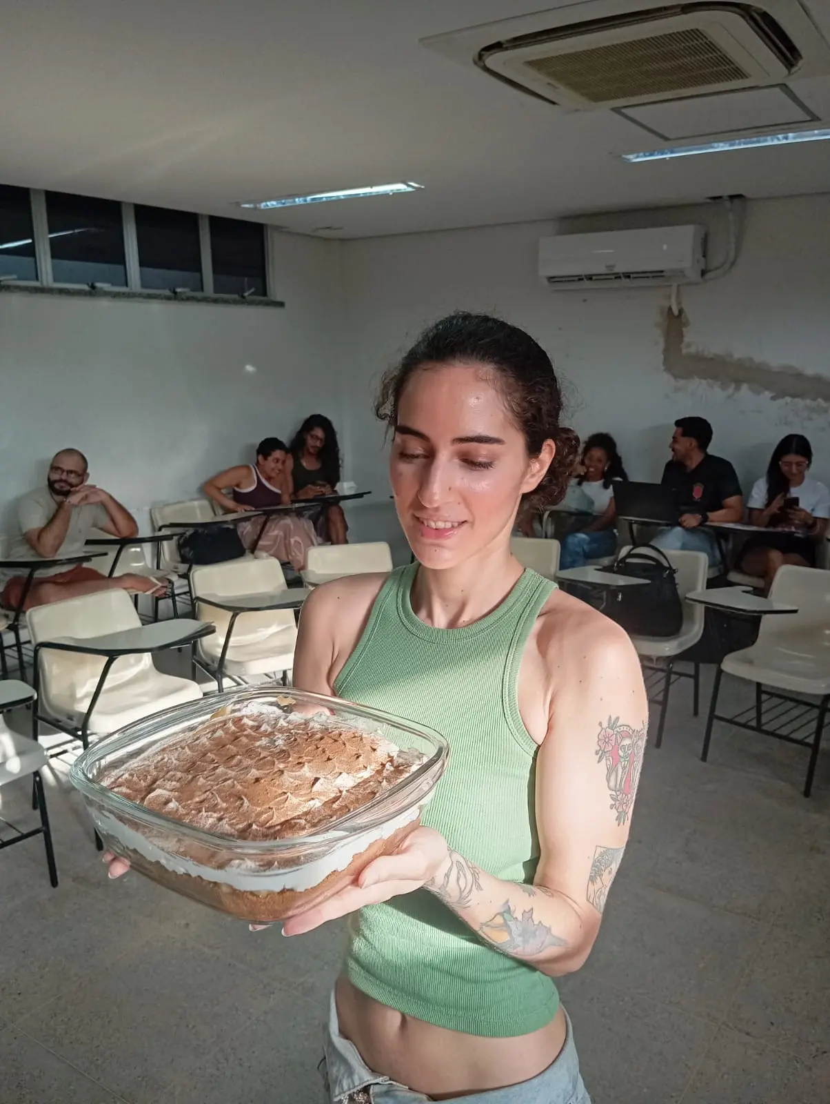 Imagem mostra uma mulher branca e jovem de camiseta verde sem mangas, com braços tatuados e cabelo castanho preso, segurando uma banoffee em uma travessa transparente e olhando para o doce com um sorriso no rosto
