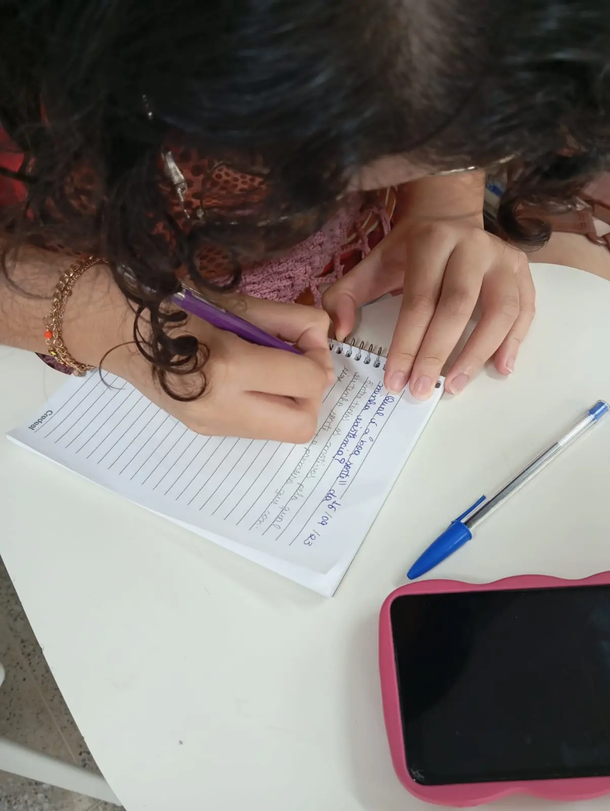 Imagem mostra mãos de uma mulher branca escrevendo com caneta roxa em caderno pequeno apoiado em uma mesa branca