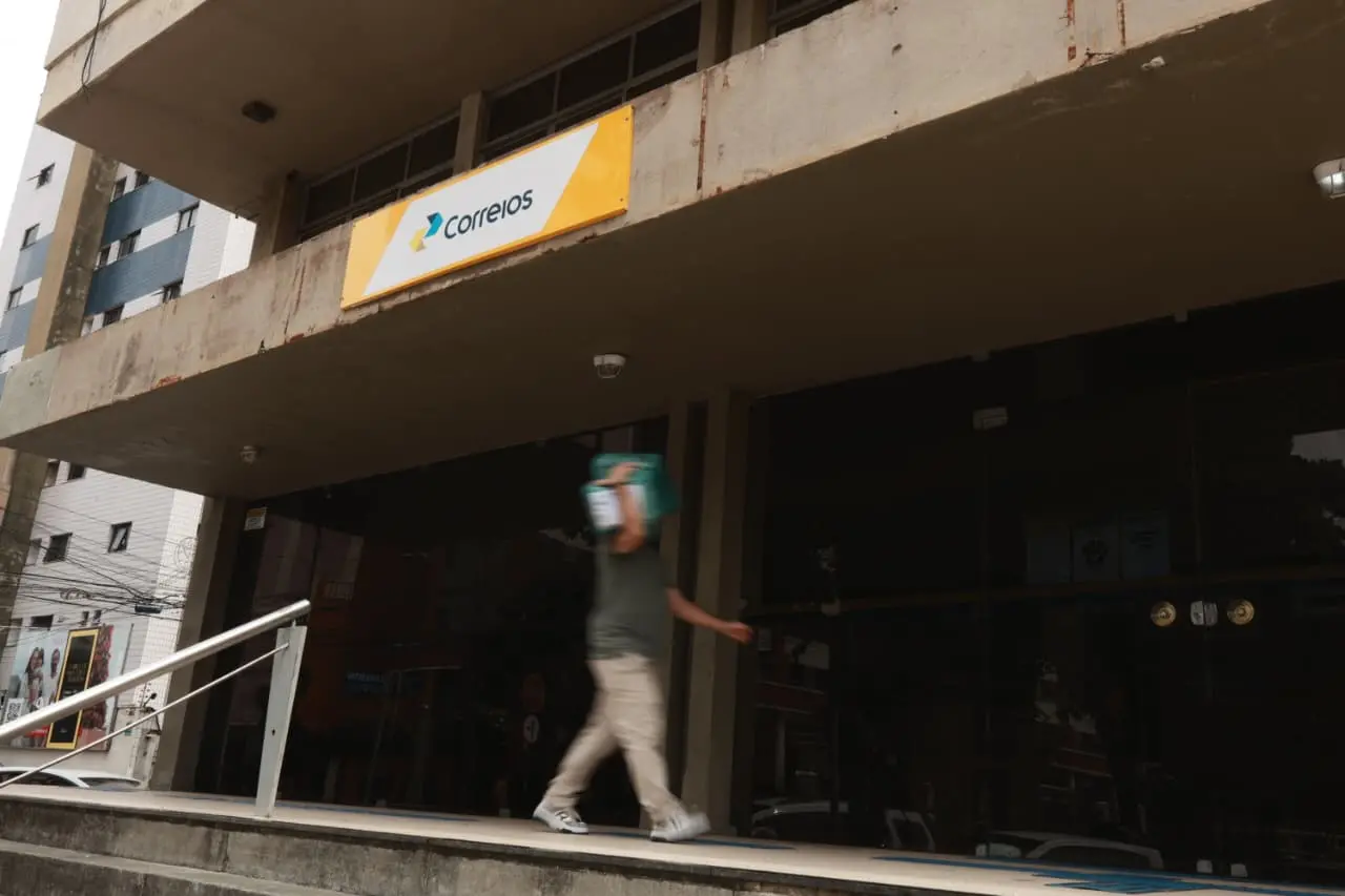 Imagem de homem carregando pacote no ombro na entrada de uma agência dos Correios.