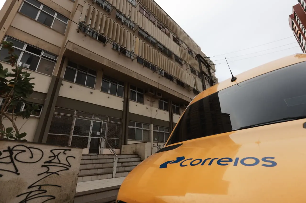 Lateral de prédio de seis andares com uma van identificada dos Correios estacionada em frente.