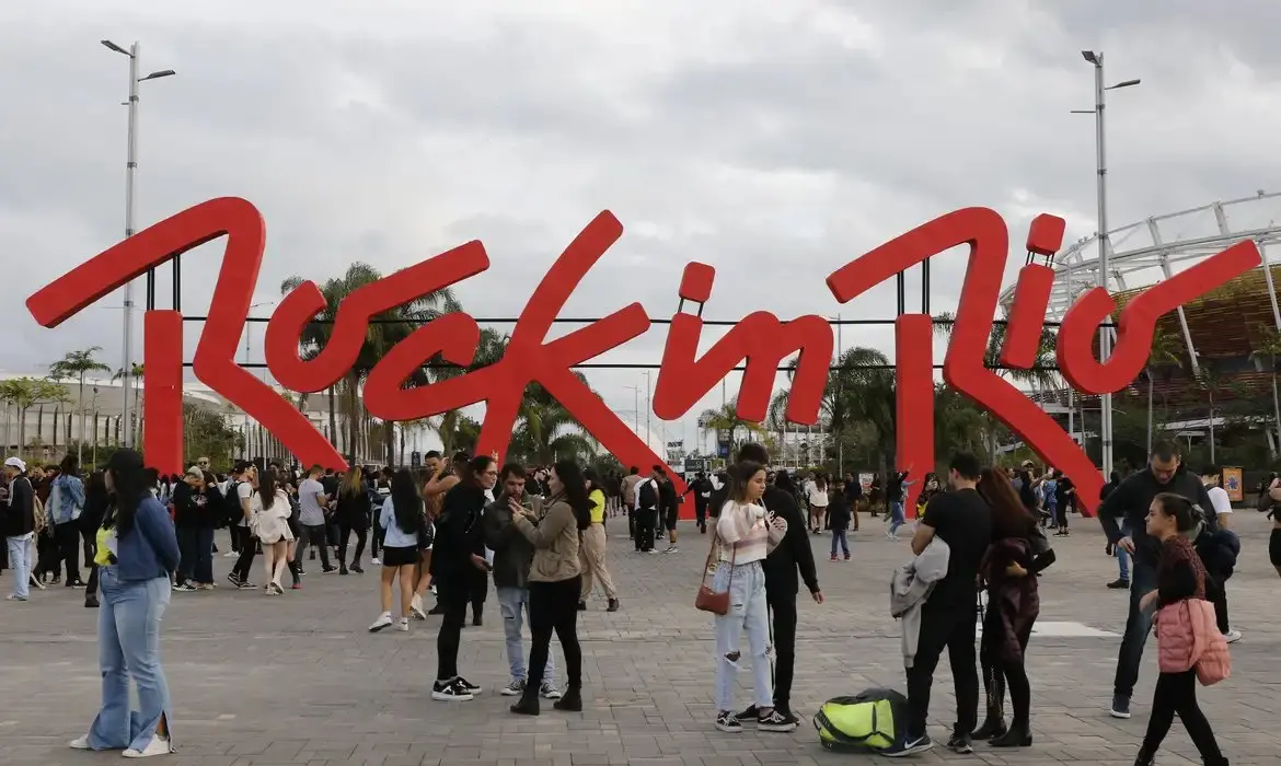 Imagem de pessoas reunidas na entrada do Rock in Rio, com o grande letreiro vermelho do evento ao fundo, em um dia nublado.