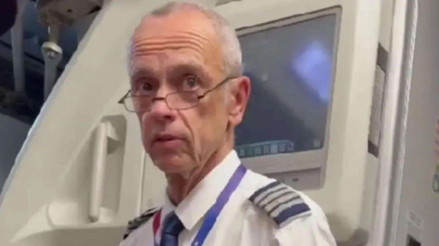 Piloto uniformizado em pé na entrada da cabine de uma aeronave, vestindo camisa branca com ombreiras de comando e crachá pendurado no pescoço; ao fundo, parte dos painéis e equipamentos internos do avião.