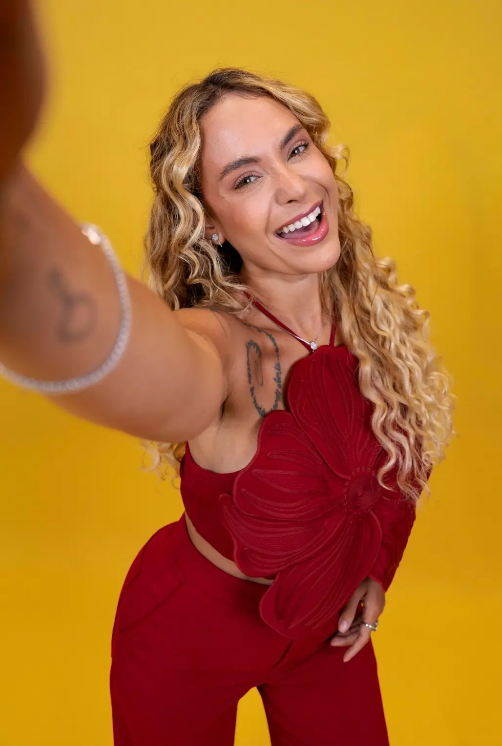 Sarah Andrade é uma mulher branca e loira de cabelo levemente cacheado. Na foto, ela usa um conjunto vermelho e simula uma selfie em frente a um fundo amarelo.