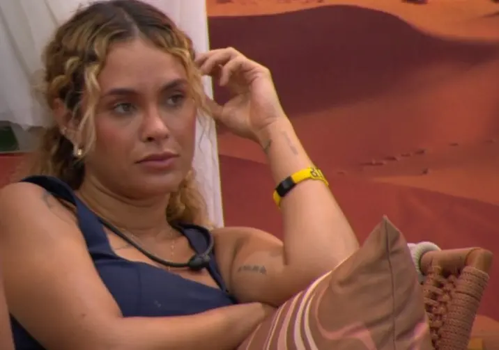 Sarah Andrade é uma mulher loira e branca de cabelo cacheado, que está preso em um rabo de cavalo. Na foto, ela está sentada e com o semblante reflexivo.