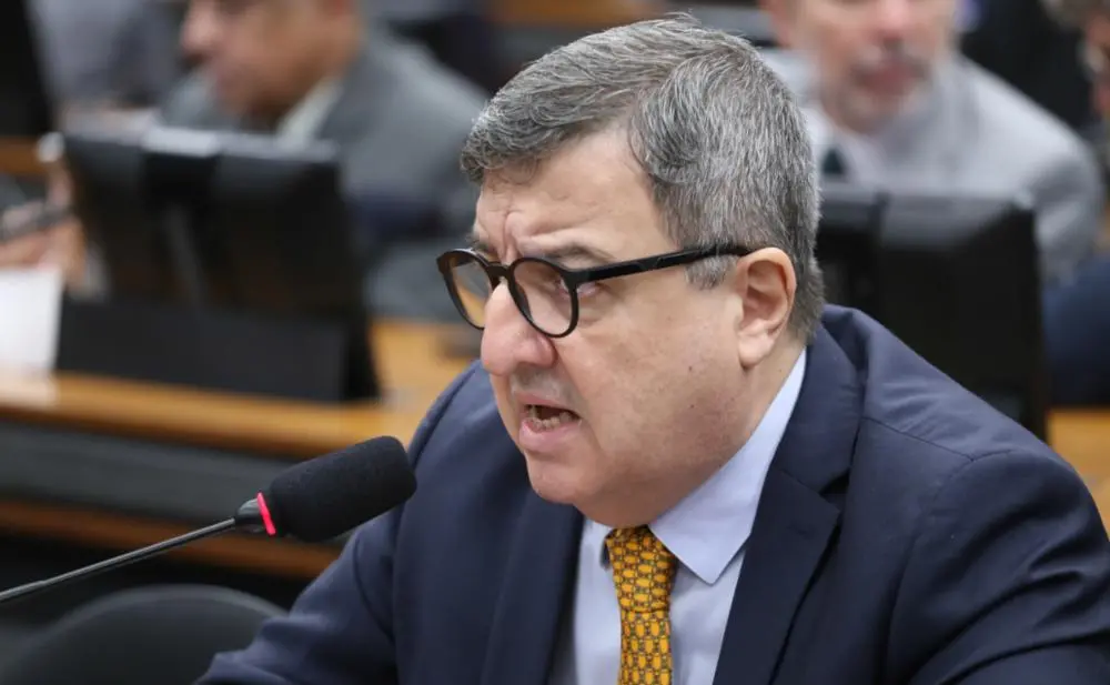 Fotografia do deputado federal Danilo Forte durante pronunciamento. Ele é um homem de pele clara, cabelos grisalhos e usa óculos de armação redonda preta. Veste terno azul, camisa azul clara e gravata amarela estampada. Ele fala ao microfone em um ambiente que remete ao plenário ou comissão da Câmara dos Deputados.