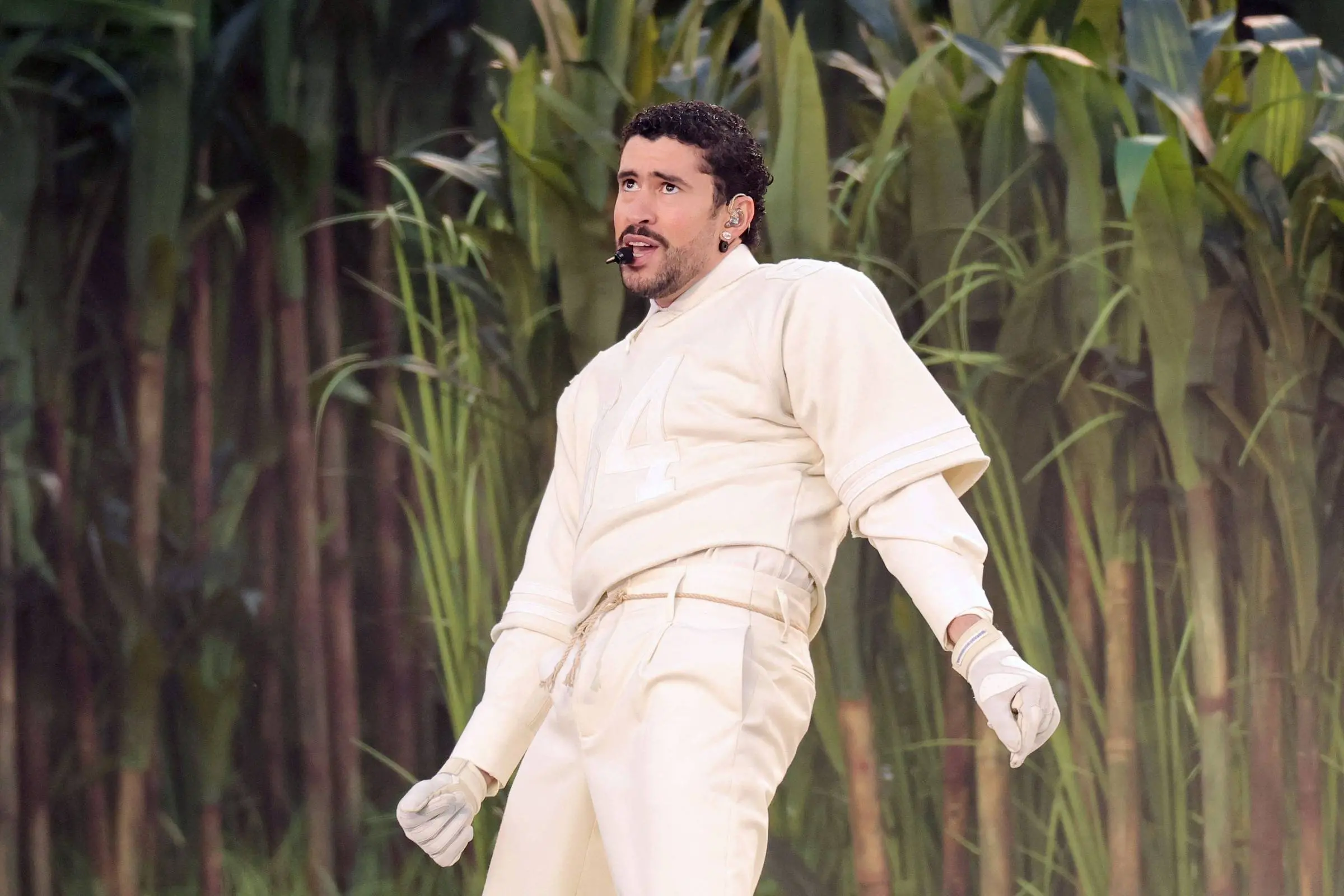Na imagem, o cantor Bad Bunny em uma performance ao vivo. Ele veste um conjunto monocromático off-white composto por uma blusa de manga longa com o número 