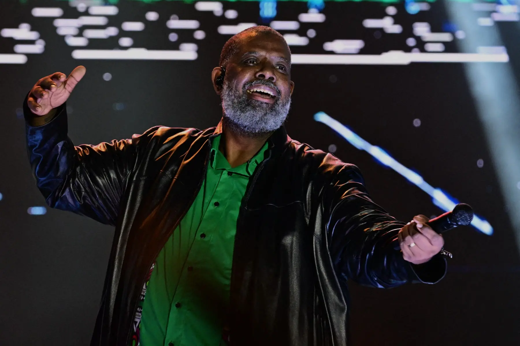 O cantor Péricles, um homem negro de barba grisalha, sorri alegremente no palco enquanto segura um microfone com a mão direita e estende a esquerda para o alto. Ele veste uma jaqueta de couro preta aberta sobre uma camisa verde vibrante. Ao fundo, um painel luminoso exibe traços e pontos brancos desregulados sobre um fundo escuro.