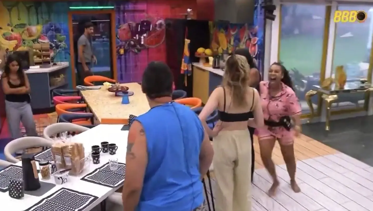 Cena de uma cozinha colorida de reality show, com várias pessoas interagindo. No centro, uma mulher de calça bege e top preto segura a mão de outra mulher vestindo pijama rosa estampado. À esquerda, uma pessoa está encostada na parede com os braços cruzados. Um homem de camiseta azul e bermuda camuflada aparece de costas, próximo à mesa posta com utensílios.