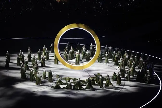 Imagem mostra artistas em trajes verdes detalhados posicionados em círculos ao redor de um grande anel dourado iluminado no centro de um palco escuro, durante uma performance na abertura da Olimpíada de Inverno 2026.