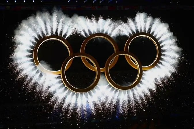 Imagem em close-up dos cinco anéis olímpicos dourados suspensos contra um fundo escuro. Os anéis estão envoltos em uma explosão simétrica de fogos de artifício brancos e faíscas que emanam de suas bordas, criando um efeito de luz radiante e fumaça clara no topo.