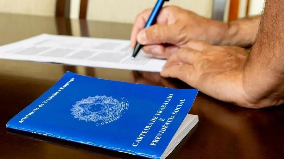 Imagem de foto em close de uma Carteira de Trabalho e Previdência Social (CTPS) brasileira sobre uma mesa de madeira escura. Ao fundo, as mãos de uma pessoa aparecem segurando uma caneta azul, prestes a assinar um documento em papel branco.