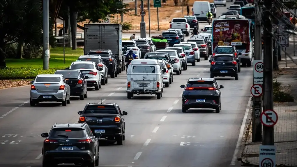 Trânsito intenso em avenida urbana, com carros, caminhões e motocicleta ocupando várias faixas; placas de sinalização indicam limite de velocidade e fiscalização eletrônica, em via arborizada durante o dia.
