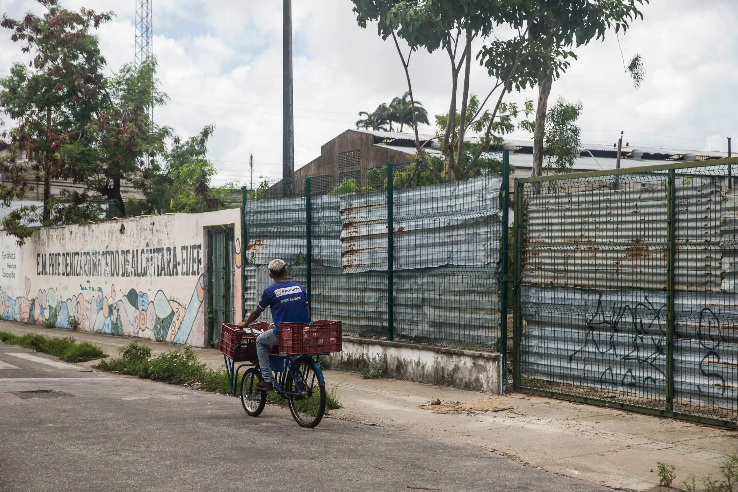 Homem em bicicleta de carga com caixas, passando por um muro colorido de escola e uma cerca metálica em ambiente urbano.