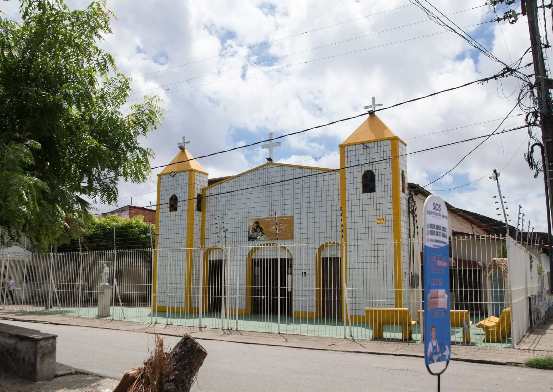 Igreja com fachada branca e detalhes em amarelo vibrante, incluindo duas torres com cruzes, vista de uma rua arborizada sob céu nublado.