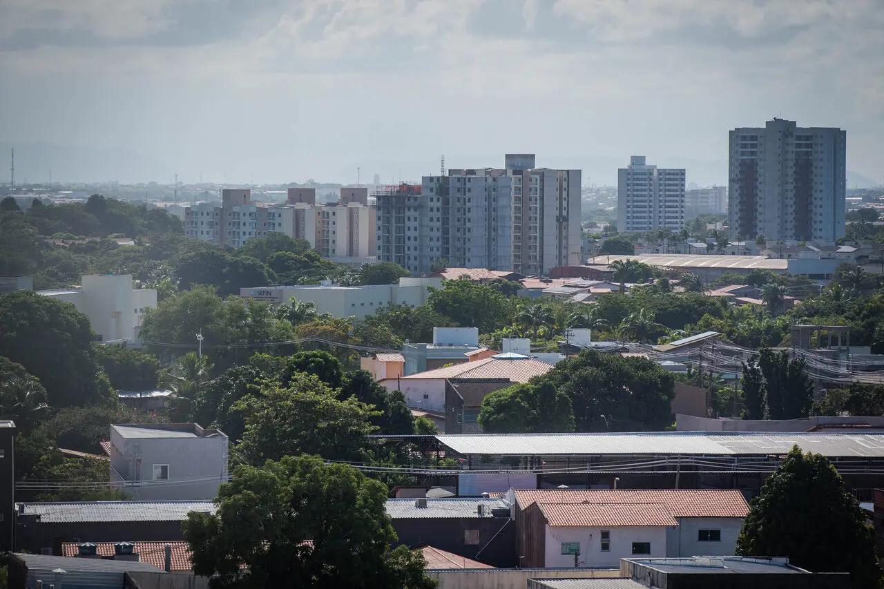 Vista panorâmica do Eusébio com prédios residenciais de diferentes alturas ao fundo e casas térreas cercadas por árvores em primeiro plano, sob céu parcialmente nublado, representando expansão imobiliária.