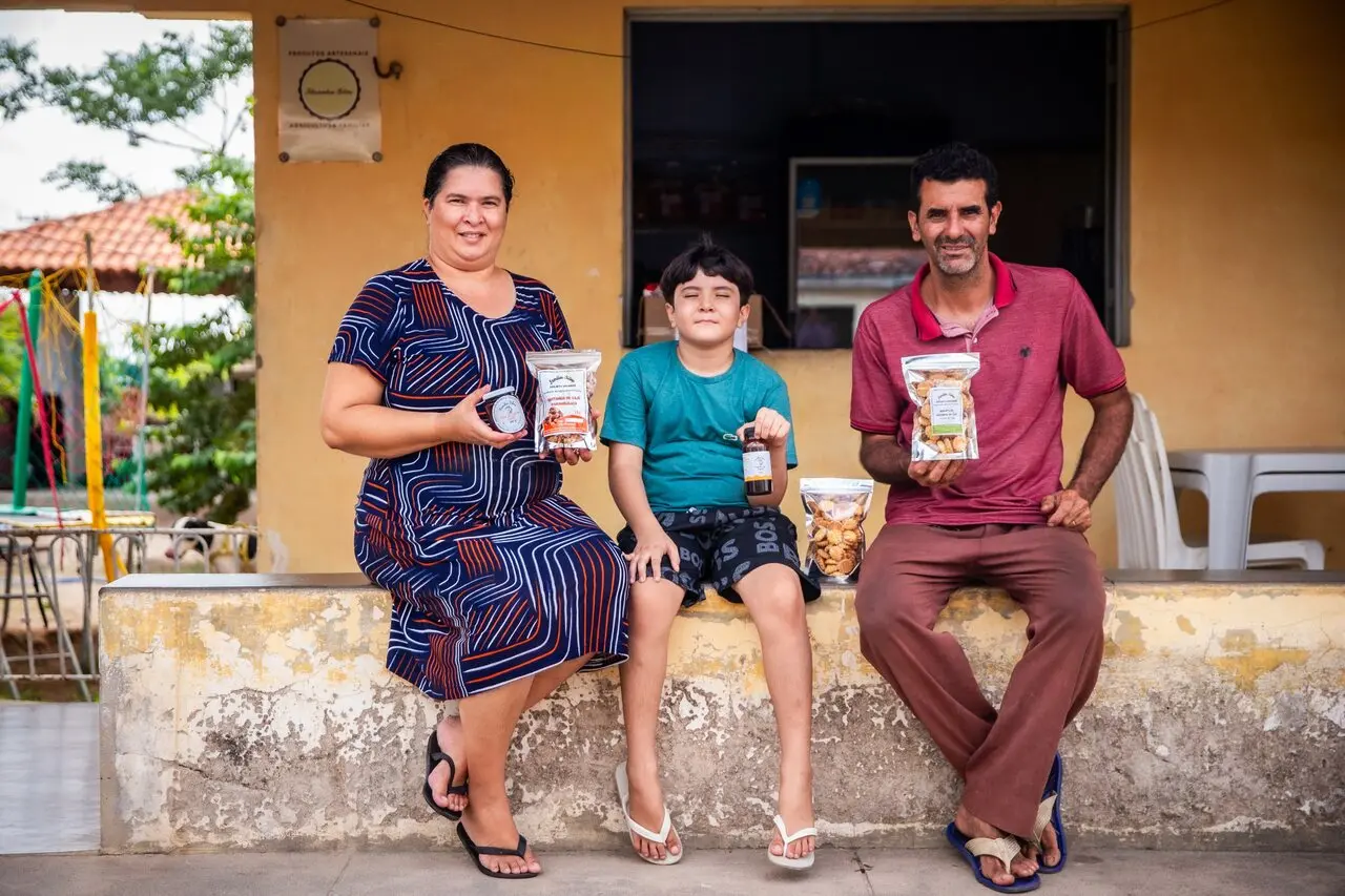 Família composta por homem, mulher e criança sentados em frente a pequeno comércio rural, segurando embalagens de castanhas e outros produtos derivados do caju, representando geração de renda no semiárido.