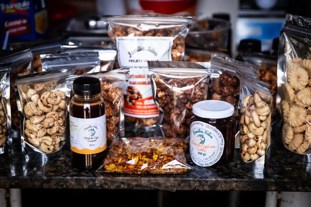 Produtos derivados do caju, como castanhas embaladas, doce em calda e mel de caju, organizados sobre bancada de cozinha, em diferentes tamanhos de embalagens transparentes.