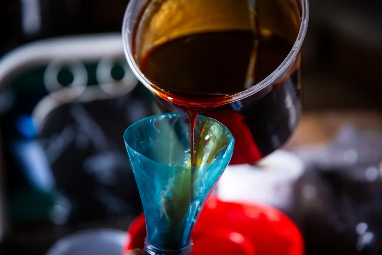 Líquido escuro à base de caju sendo despejado de recipiente plástico em funil azul, em etapa de preparo artesanal de bebida ou doce.