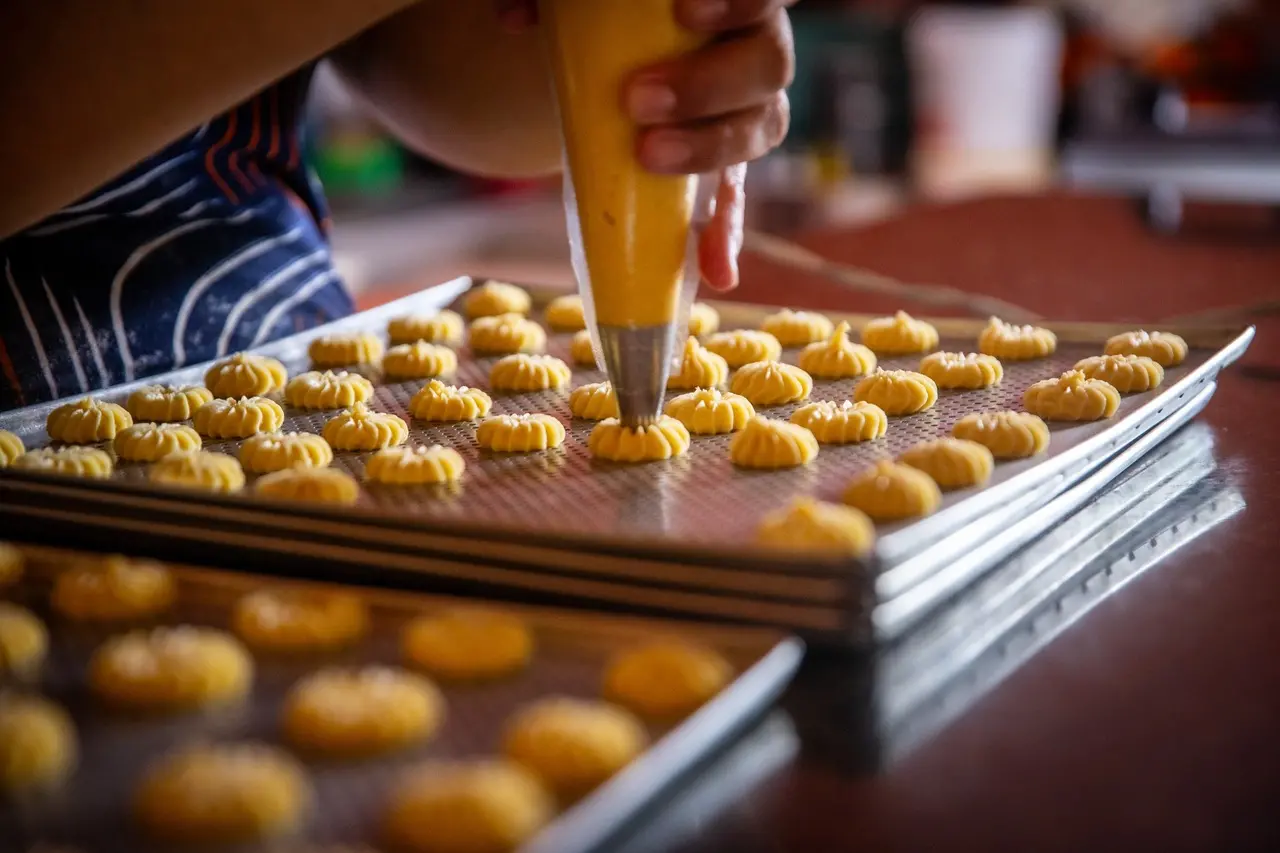 Pessoa utiliza saco de confeitar para moldar pequenos biscoitos sobre assadeiras metálicas, em processo artesanal de produção de alimentos.