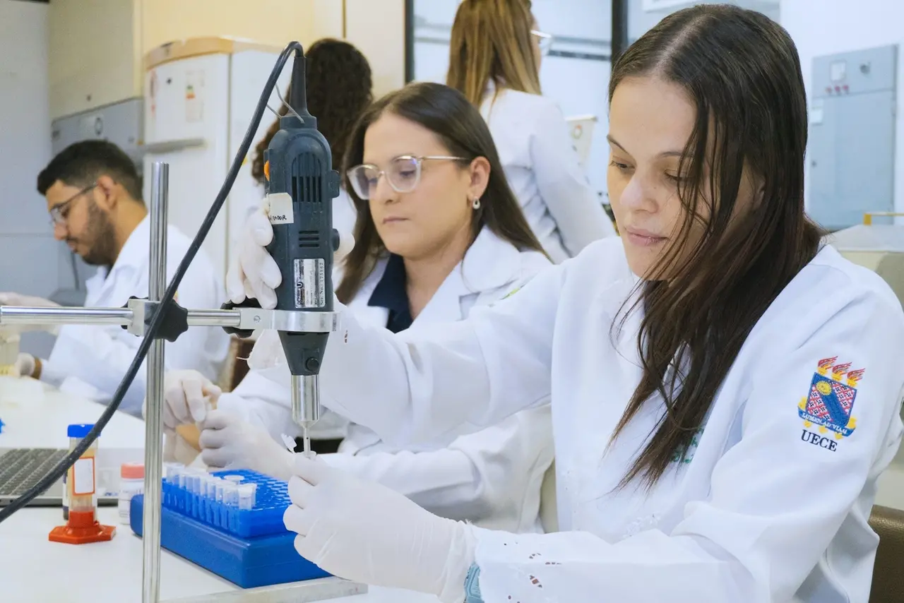 Imagem mostra jovem pesquisadora realizando experimentos laboratoriais, usando jalecos brancos, em um ambiente de curso técnico ou faculdade.