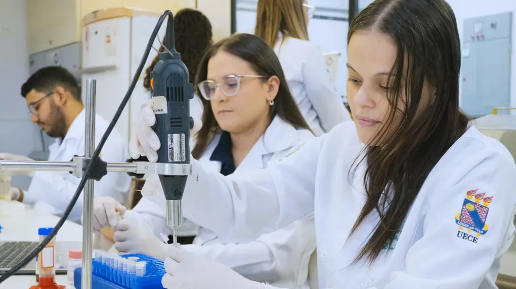 Imagem mostra jovem pesquisadora realizando experimentos laboratoriais, usando jalecos brancos, em um ambiente de curso técnico ou faculdade.