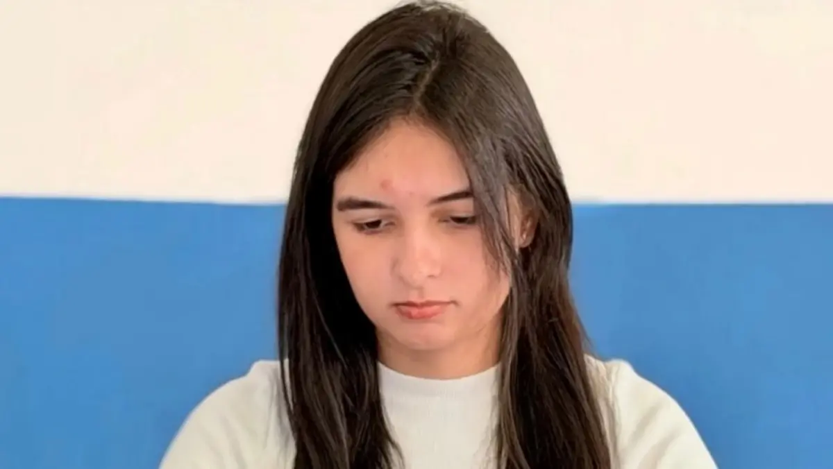 Fotografia de plano médio da estudante de psicologia Vanessa Lara. Ela tem cabelos castanhos longos e lisos que caem sobre os ombros e veste uma blusa branca de gola redonda. Vanessa está com o rosto levemente inclinado para baixo, com uma expressão séria e concentrada. O fundo é composto por uma parede dividida horizontalmente nas cores branco e azul.